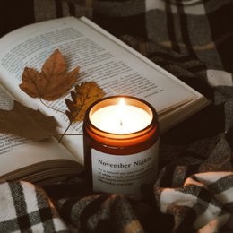 A round, tall candle in a brown glass container labeled "November Nights 155 g. Candle". The candle is lit, casting a warm glow, and is surrounded by autumn leaves on an open book.