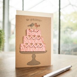 A pink wedding cake with hearts on a pedestal, depicted on a greeting card with the text "We get hitched!". The card is made of brown kraft paper, giving it a rustic, handmade feel.