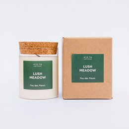 A tall, round green candle with a cork lid, surrounded by fresh citrus fruits and greenery. The candle is labeled "Lush Meadow" and appears to be a scented candle product.
