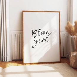 Decorative poster featuring the text "Bloom girl" in a minimalist, handwritten style. The poster is displayed in a simple wooden frame against a white wall and wooden floor, creating a cozy and feminine atmosphere.