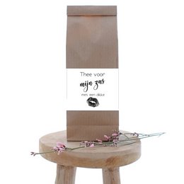 A brown paper bag with the text "Thee voor mijn zus" (Tea for my sister) printed on it, along with a small decorative item placed on a wooden surface.