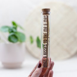 A glass tube filled with dried flowers, leaves, and other natural elements. The tube has a cork stopper and a decorative label that reads "Hugs & kisses to the mr & mrs".