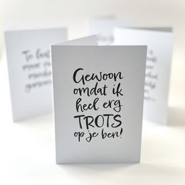 Folded A6 card with envelope, featuring handwritten text in black ink that says "Gelukkig andatik heel erg TROTS op je bent!" which translates to "Happy that I'm very PROUD of you!" The card has a simple, minimalist design with a white background.