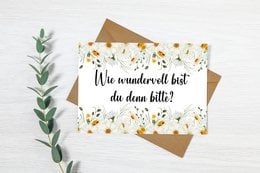 Floral postcard with handwritten text in German "Was wundervolles hast du denn bitte?" (What wonderful thing do you have, please?). The postcard is surrounded by green leaves and a brown envelope.