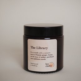 A round, tall, black candle in a glass container labeled "The Library 100 g. Kerze". The candle is a scented candle, with a warm, cozy fragrance.